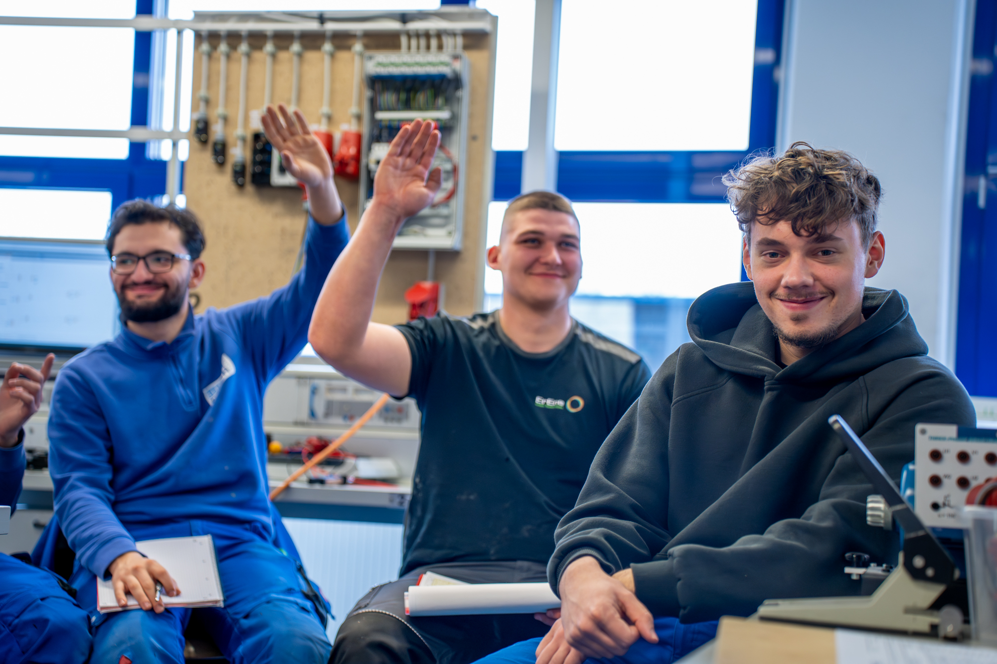 Zwei junge Männer in einer Elektrowerkstatt, einer hebt die Hand und lacht, während der andere entspannt in die Kamera schaut – Ausbildung bei der Kreishandwerkerschaft Mettmann.